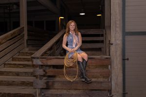 Young woman in western outfit holding a rope lasso on barn steps in cowboy boots.