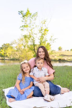 Mother smiling with young daughter in blue dress and toddler son on outdoor blanket.