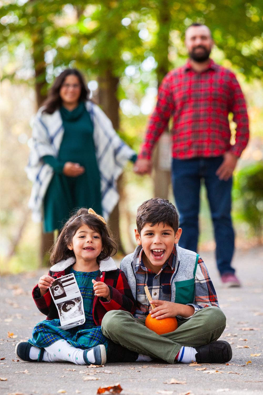 Two excited children sitting on a path holding a sonogram and pumpkin with parents standing behind.