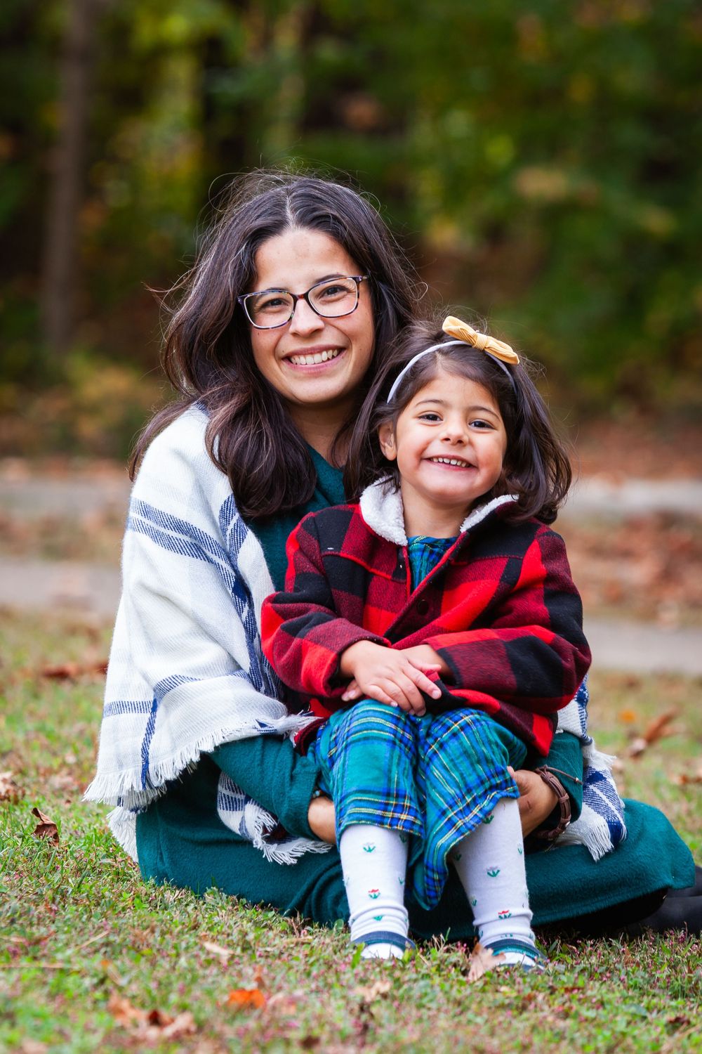 A smiling mother with glasses and her young daughter sit together outdoors in autumn, both wearing plaid and cozy layers.