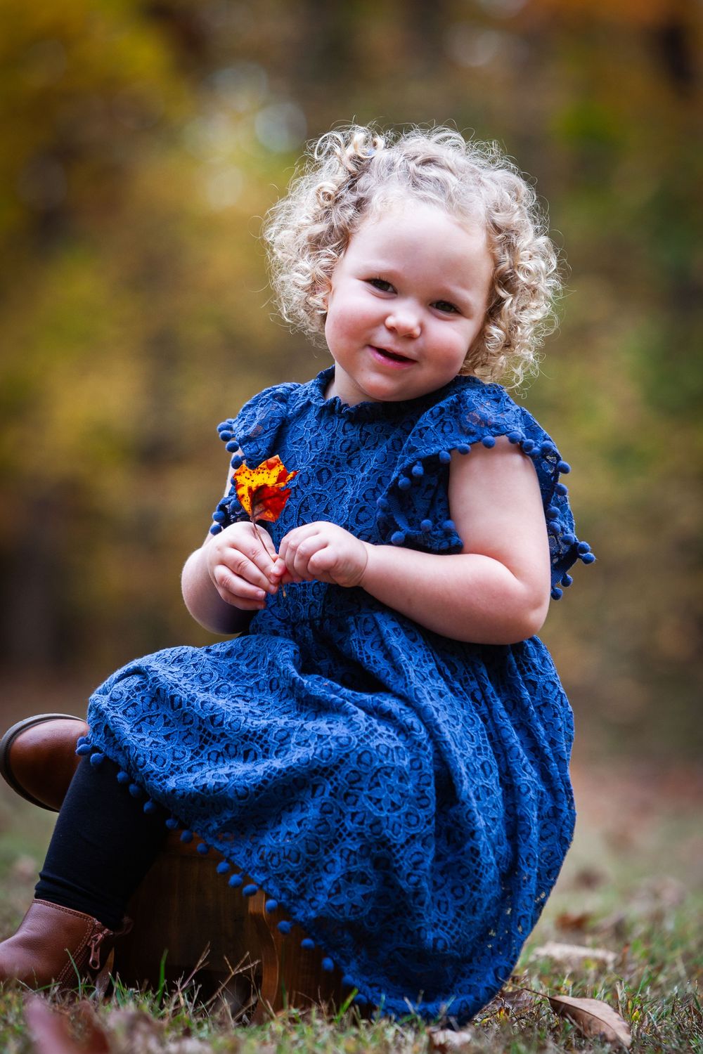 Curly-haired toddler girl in blue dress holding orange flower, smiling outdoors in autumn setting.