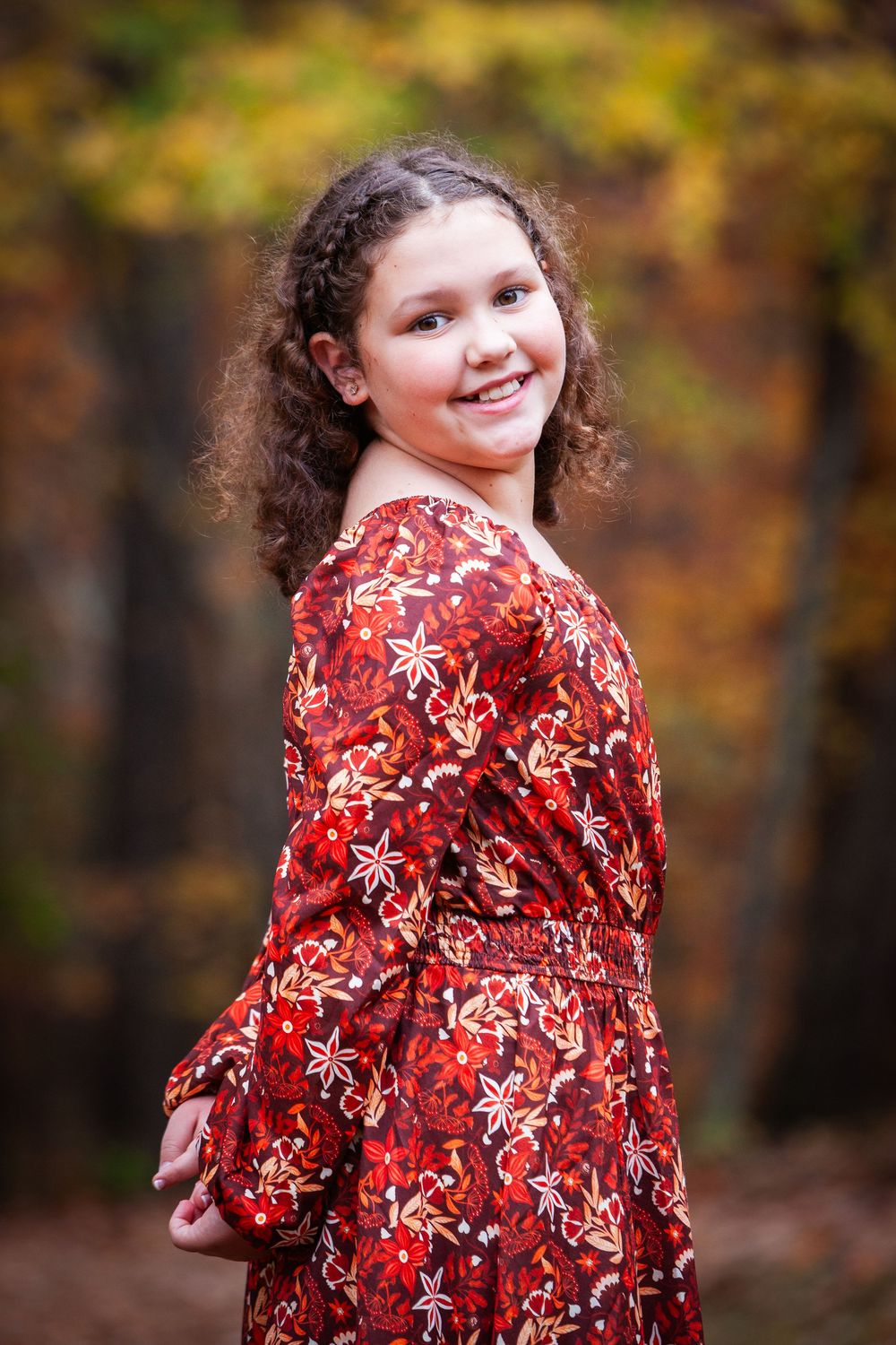 A smiling young girl with curly hair wearing a red floral dress poses outdoors in an autumn forest setting.