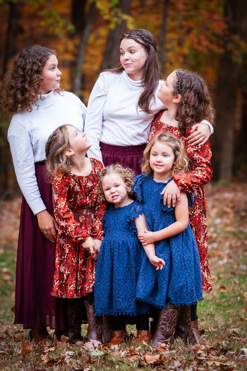 Six girls in autumn dresses pose together outdoors among colorful fall foliage for a family portrait.