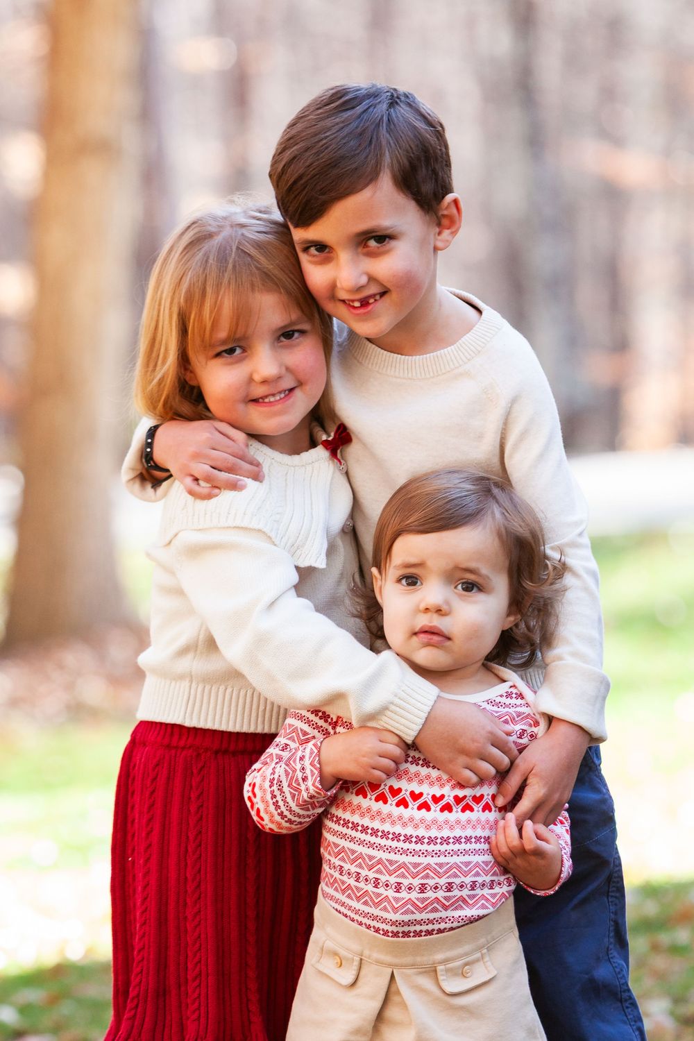 Three young siblings hugging each other warmly in cream and red sweaters in an autumn park setting.