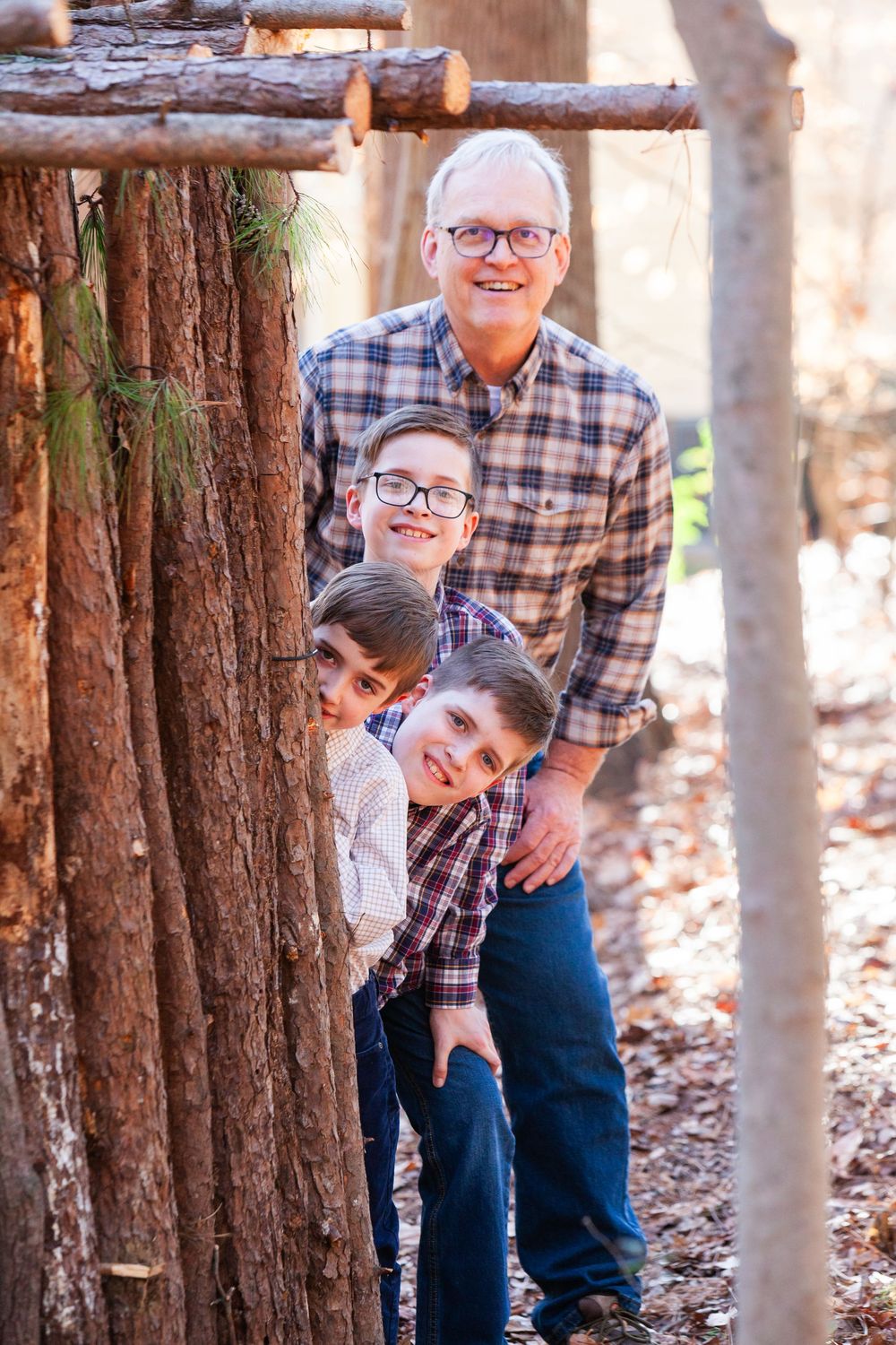 Grandfather and three grandsons smiling and peeking around pine trees in a sunny woodland setting.