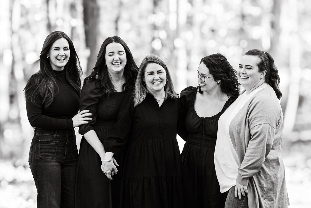 Black and white portrait of five women smiling and holding hands together in a wooded outdoor setting.