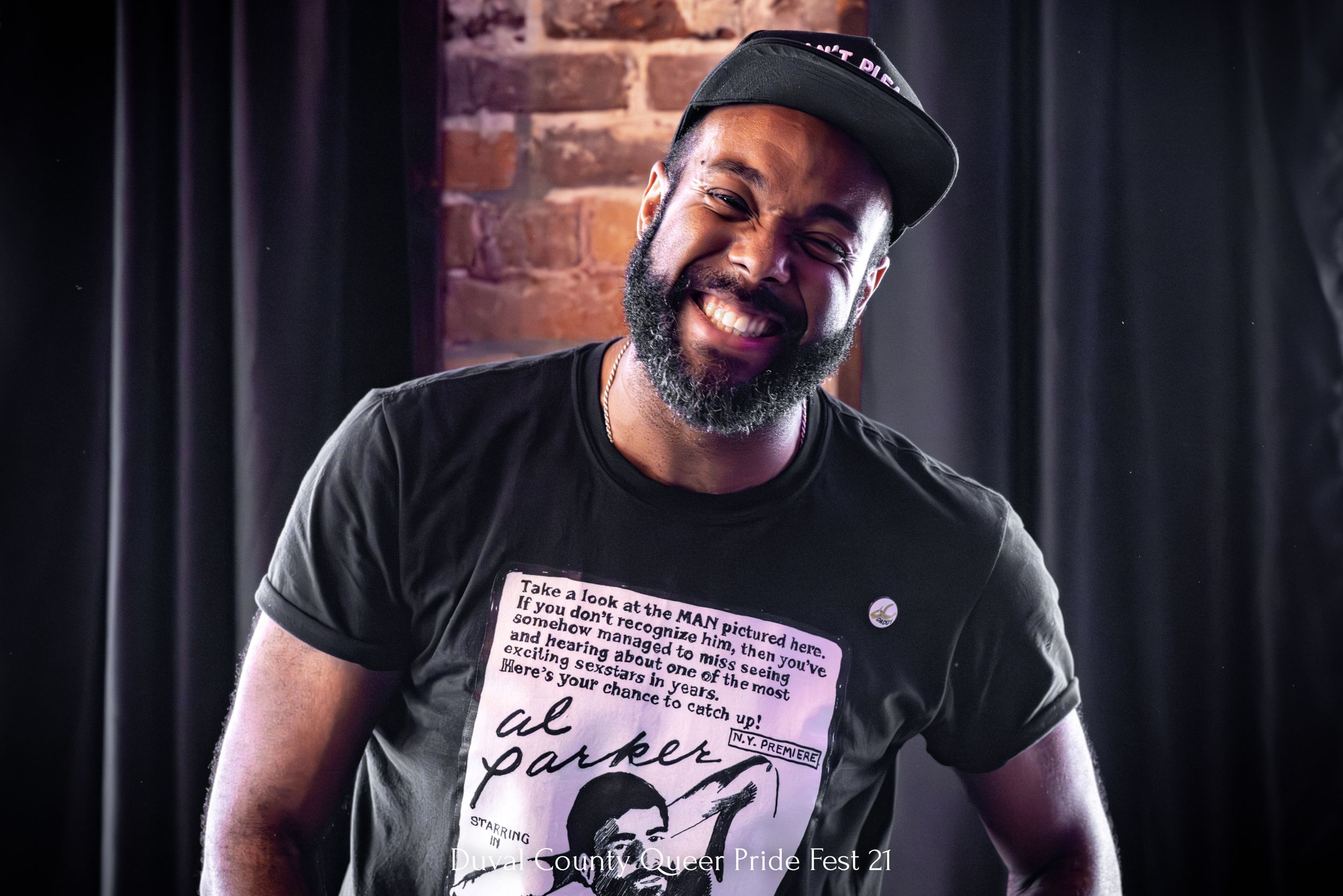 A smiling person wearing a black t-shirt and baseball cap against a dark background.