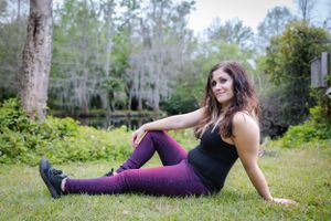 Woman in purple pants and black top sitting casually on grass with trees in background.