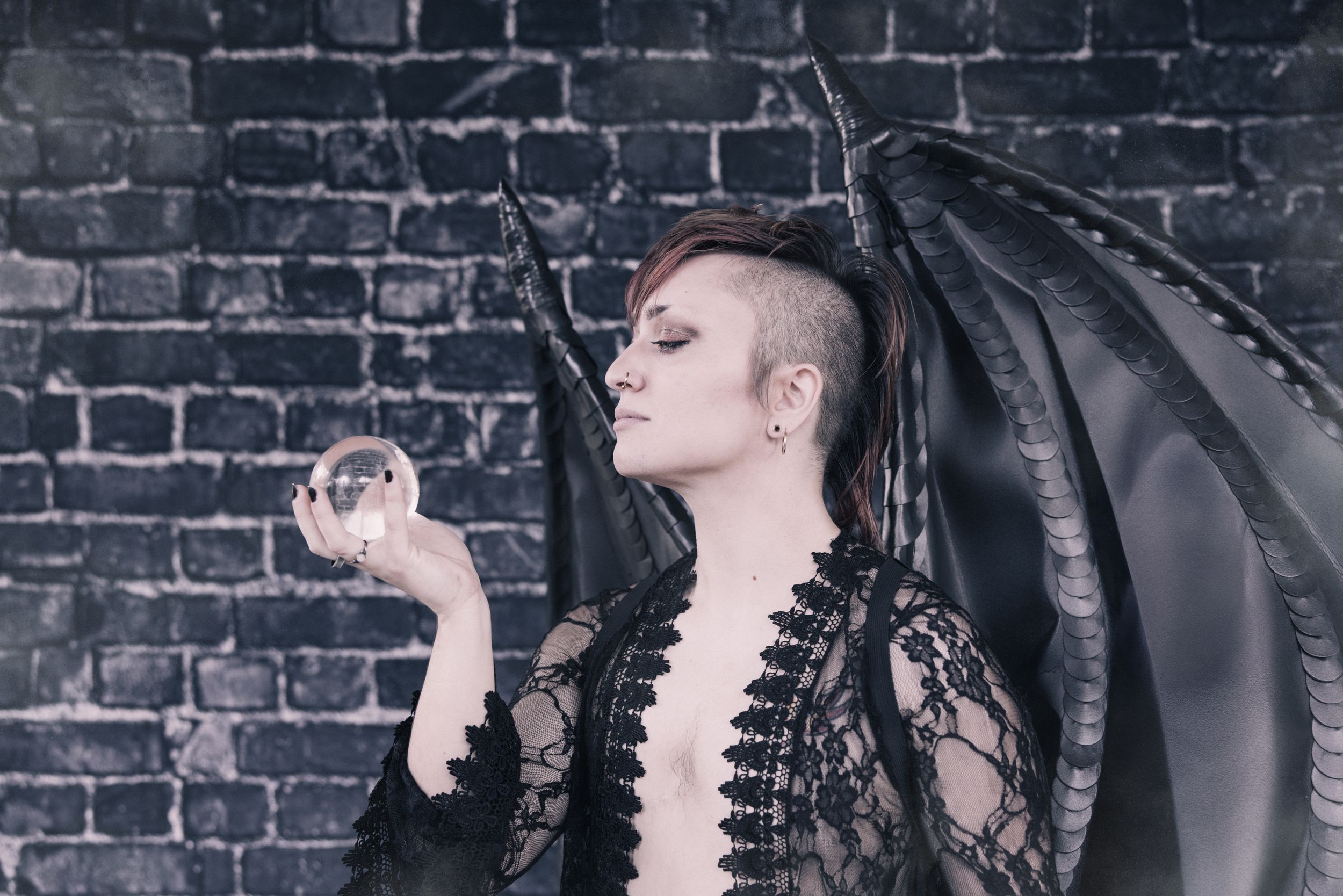 A gothic-styled figure with black wings poses against a brick wall.