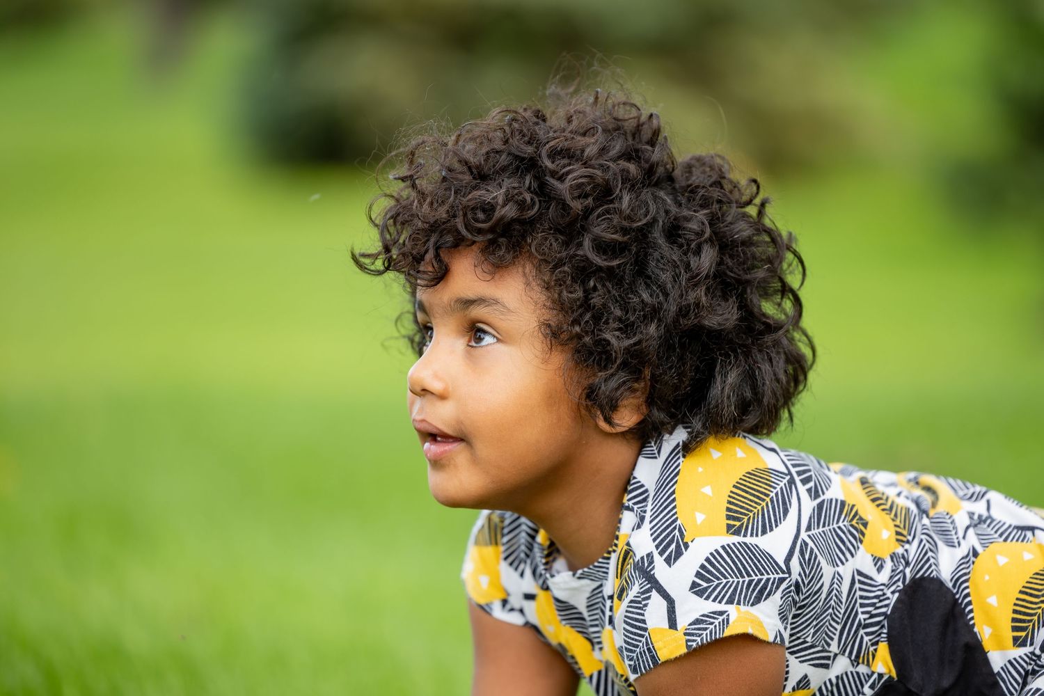Child with curly hair looks to the side while wearing a patterned shirt outdoors.