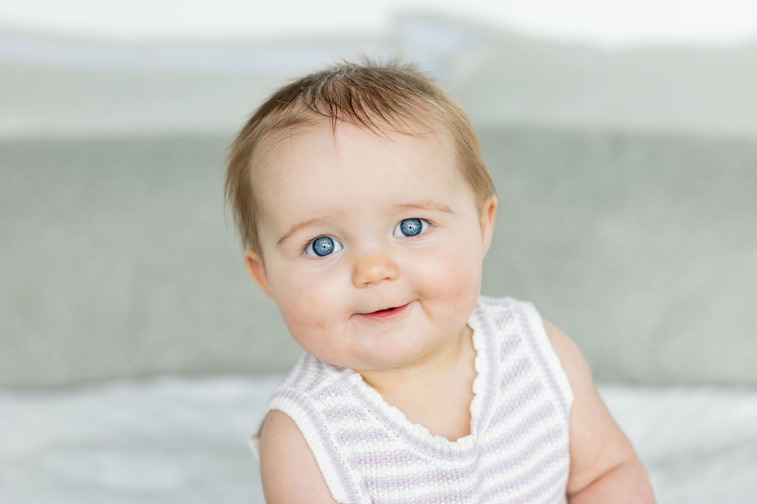 Adorable baby with blue eyes and brown hair smiling, wearing a striped sleeveless outfit, sitting on a light gray bed.