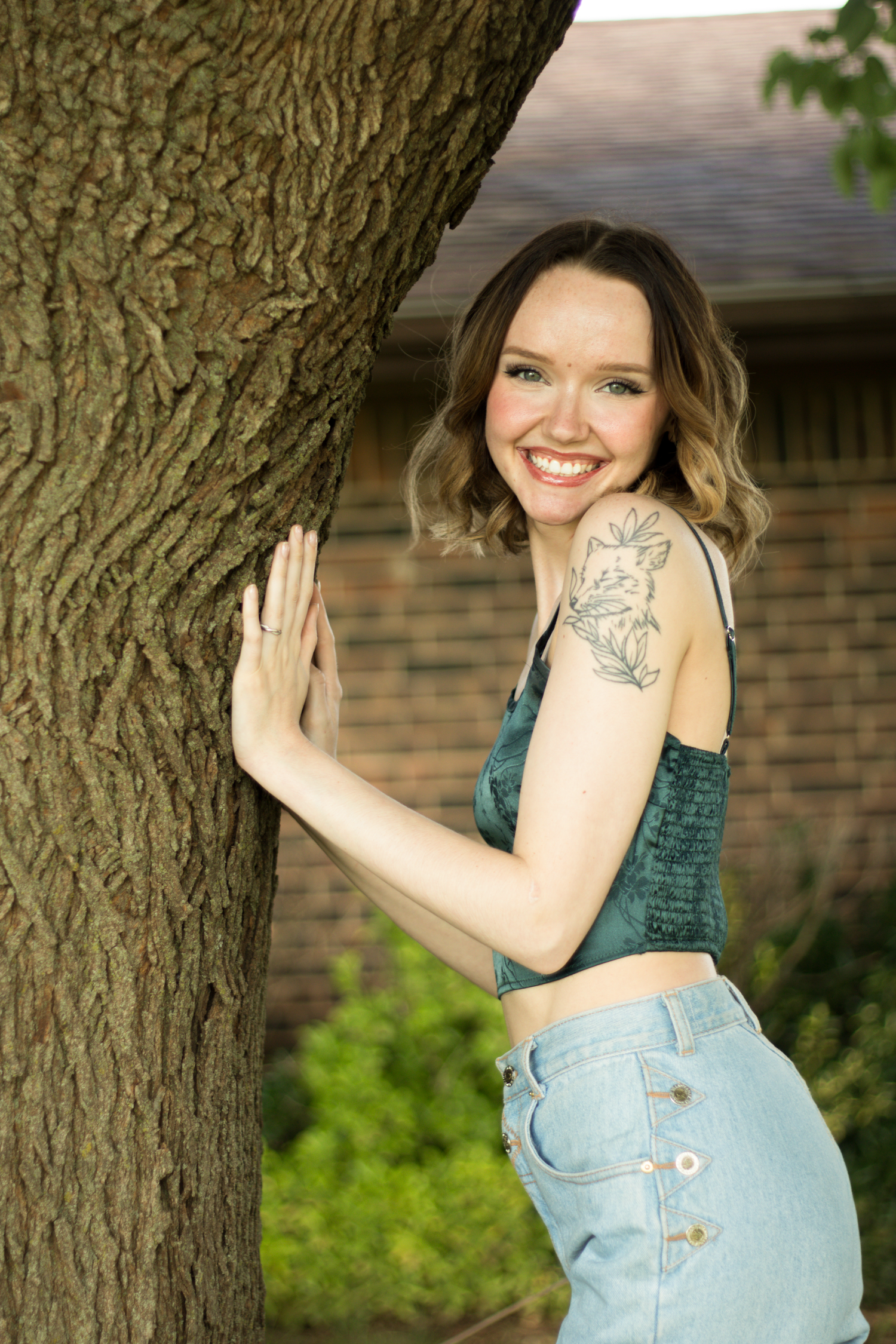 Smiling young woman with shoulder tattoo in green crop top and jeans leaning against a tree outdoors.