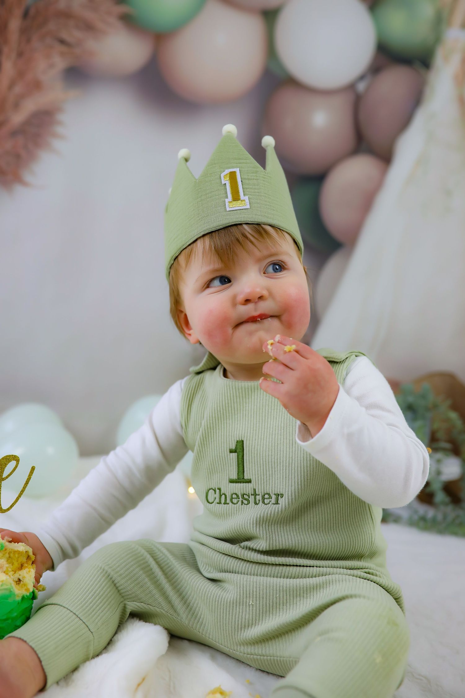 A small baby wearing a sage green outfit with matching crown hat enjoys their first birthday celebration.