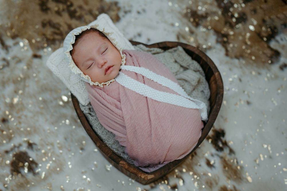 A sleeping newborn baby swaddled in pink fabric lies peacefully in a heart-shaped wooden bowl.
