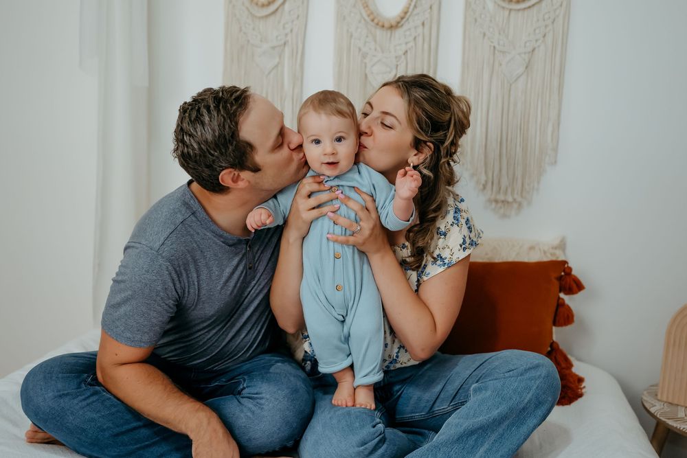 Family portrait session showing joyful moments and kisses with baby in light blue outfit against white wall.