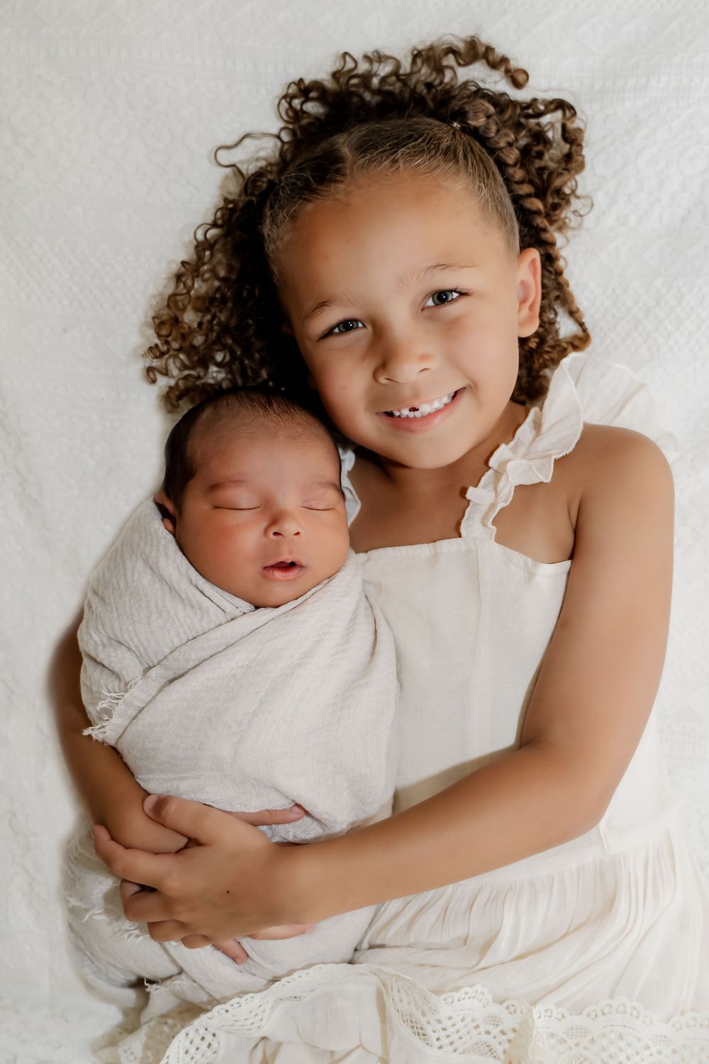 A tender series of portrait photos showing a young child holding and embracing a swaddled newborn on white bedding.