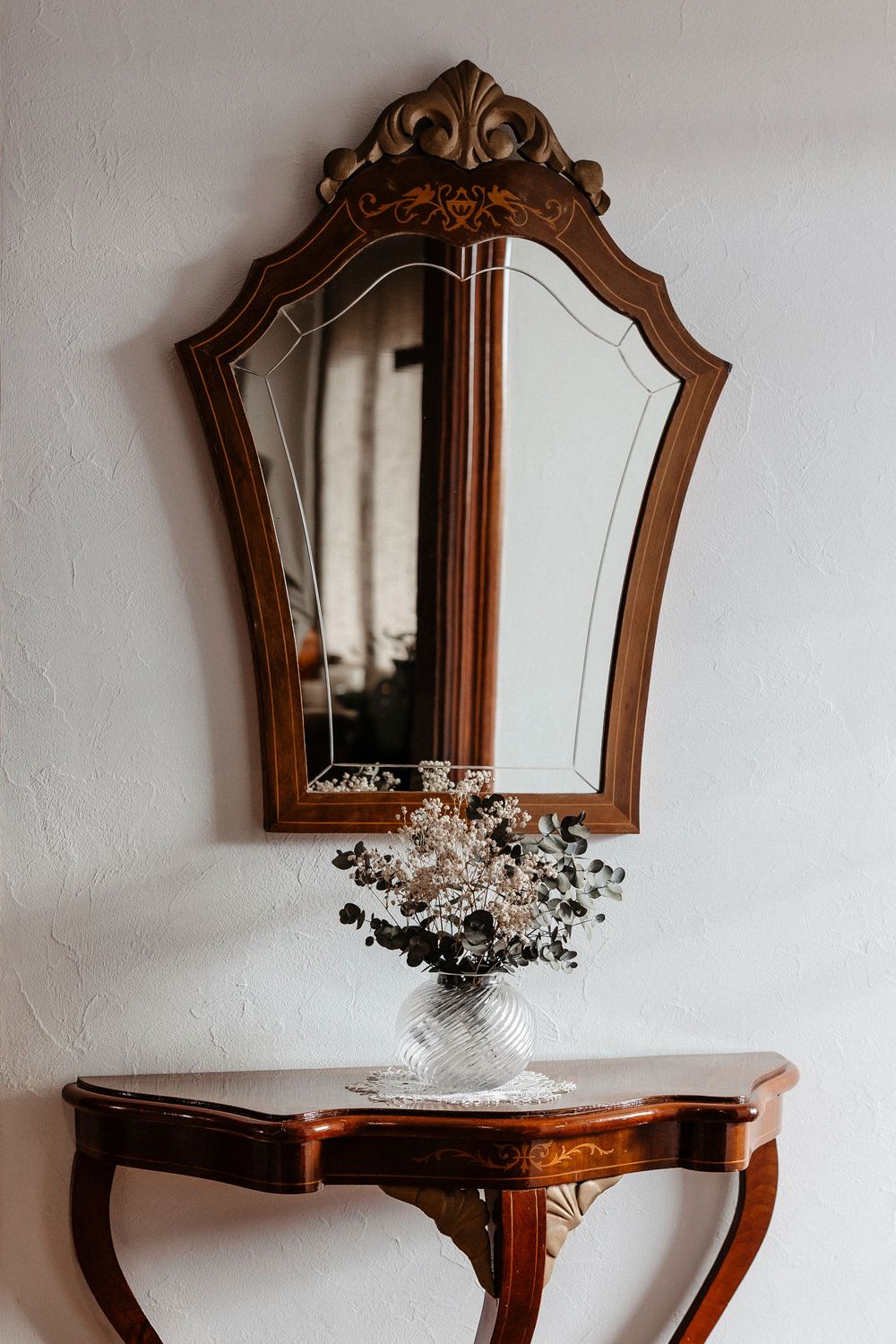 Vintage wooden console table with ornate mirror and white floral arrangement in vase against textured white wall.