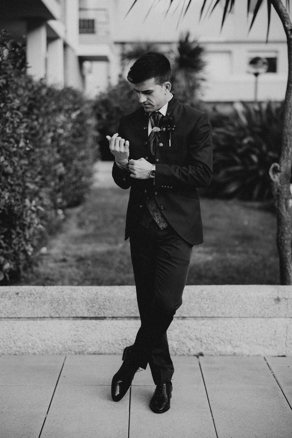 Black and white photo of person in formal suit checking watch while standing on outdoor patio.