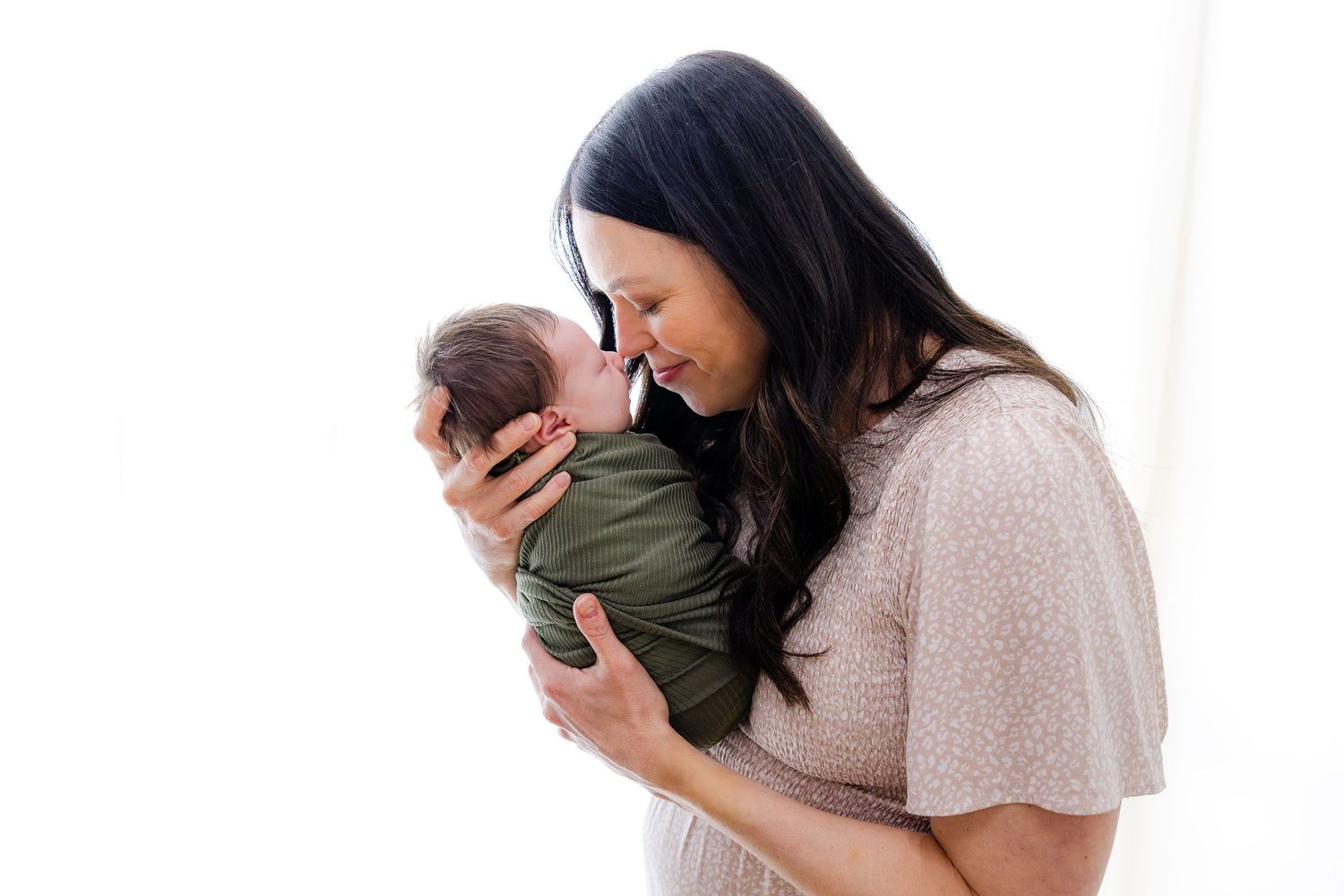 A smiling woman in a teal sweater holds a newborn baby gently in her arms, both set against a softly blurred home.