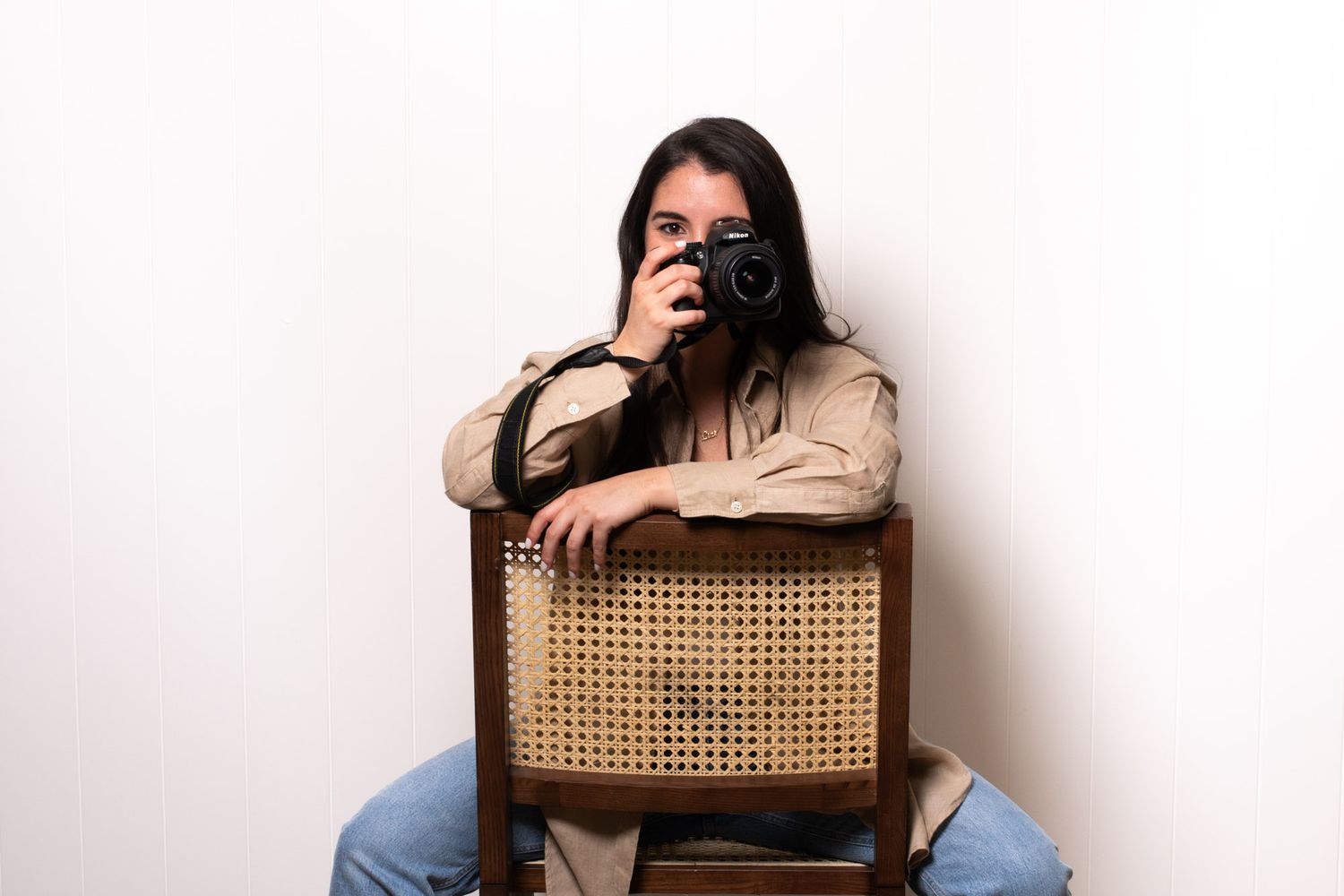 Photographer sitting on rattan chair holding camera against white wall.