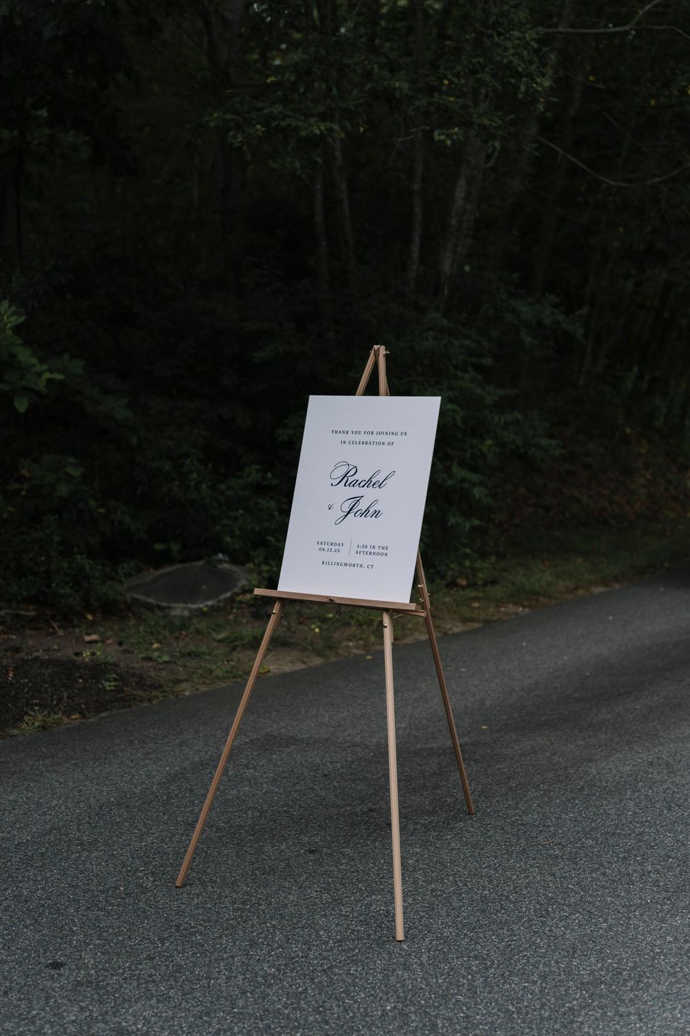 Wooden easel with white sign stands on gravel path at edge of wooded area.