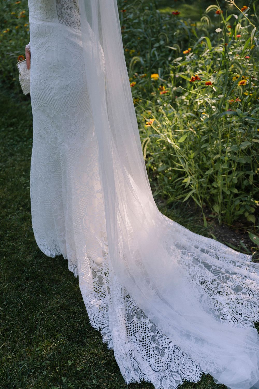 Delicate lace details of a flowing white wedding dress train against green grass and garden flowers.