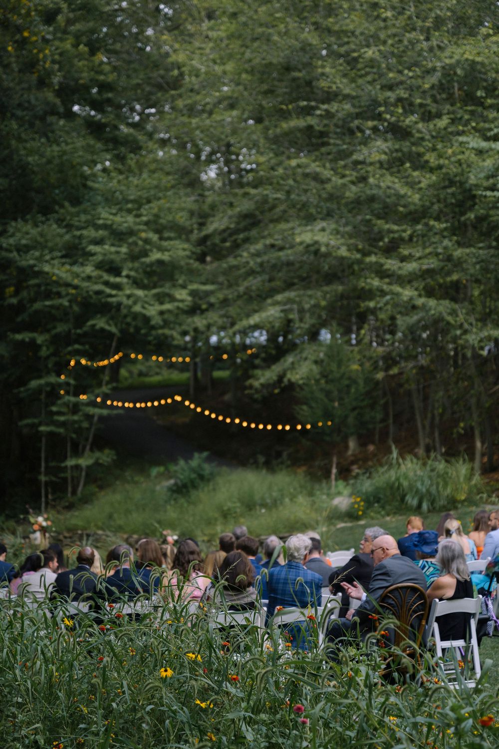 Outdoor wedding ceremony setup with string lights and chairs surrounded by trees at dusk.