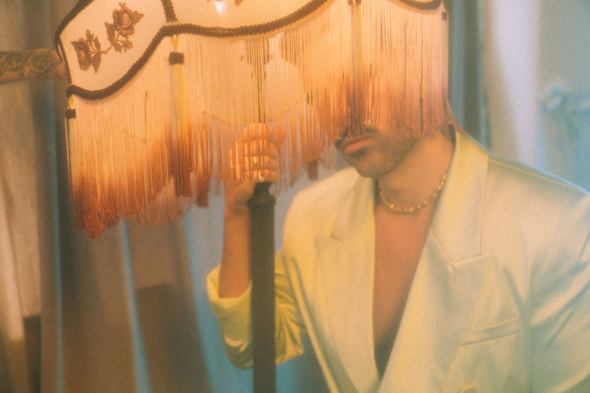 Vintage-style photo showing a person in a cream blazer and yellow shirt under warm lighting with a fringed lampshade.