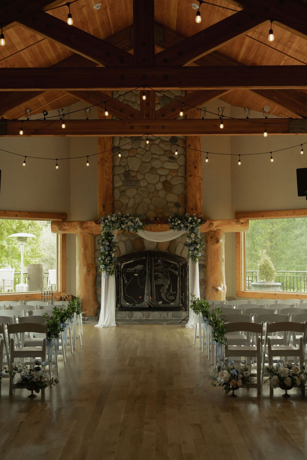 Rustic wedding venue features exposed wooden beams, string lights, and a stone fireplace altar.