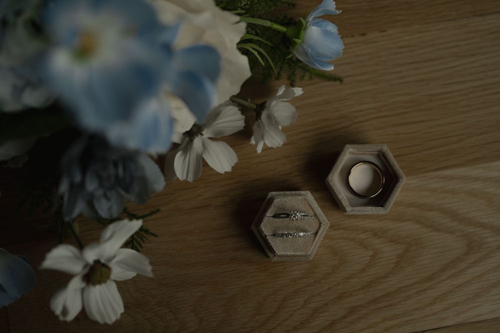 Wedding rings displayed in hexagonal boxes beside blue and white flowers on wooden surface.