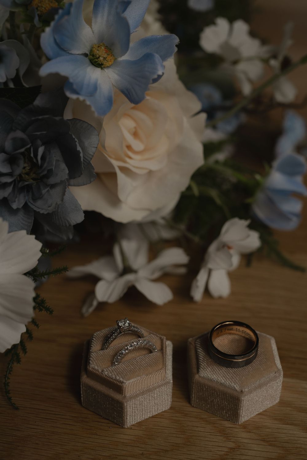 Elegant floral arrangement with blue and white blooms next to wedding rings in hexagonal holders.