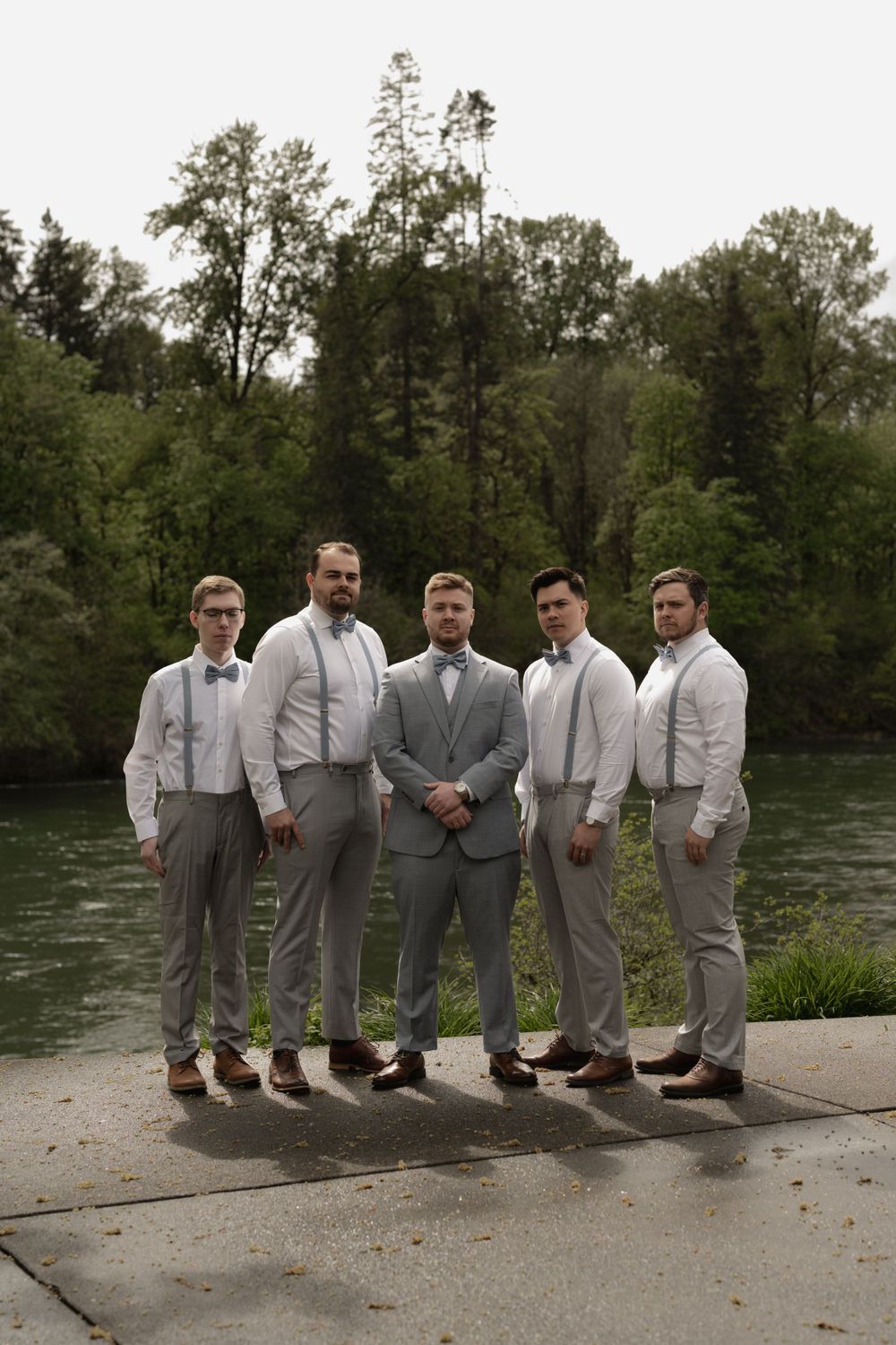 Groomsmen in coordinated gray and white attire posed by river.