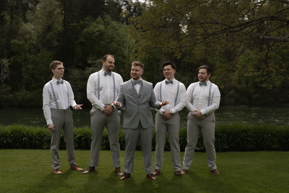 Wedding party in gray suits standing on grass with trees in background.