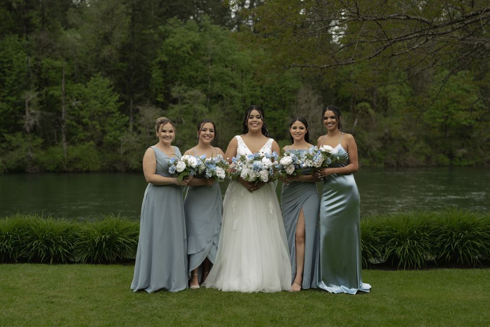 Wedding party poses by a lake with bridesmaids in blue dresses holding white bouquets.