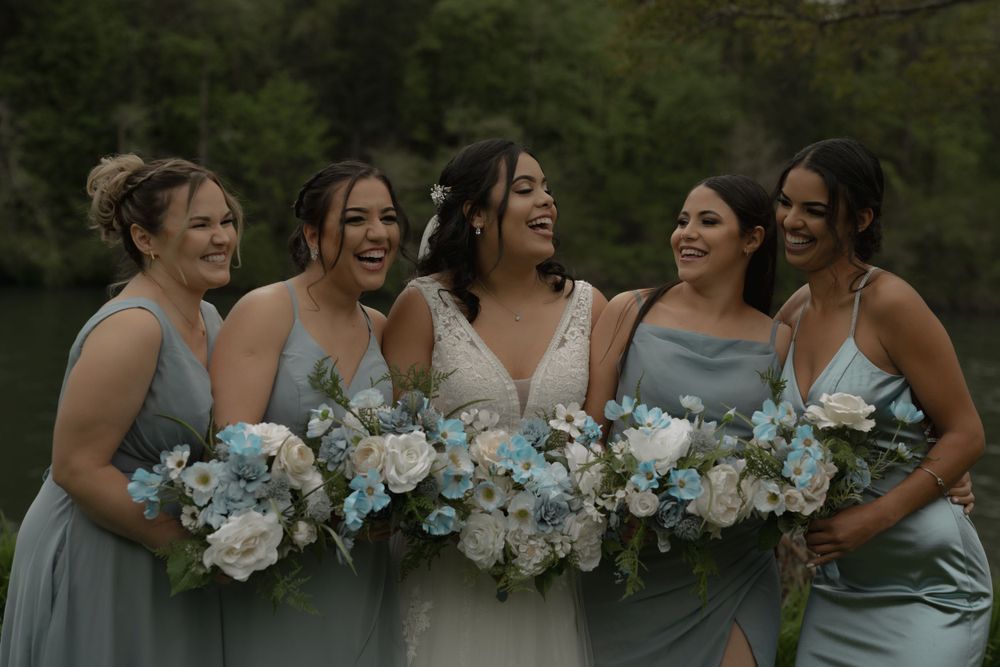 Bridesmaids in light blue dresses share a candid moment while holding floral bouquets.