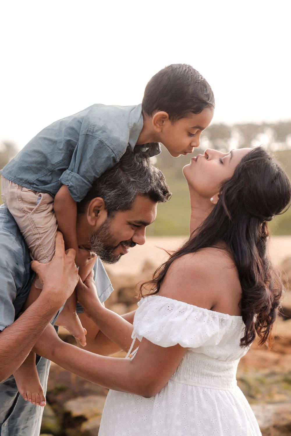Pregnant woman in a flowing white dress kissing her son while husband has the son on his shoulders at Little Bay Beach, Sydney by the ocean.