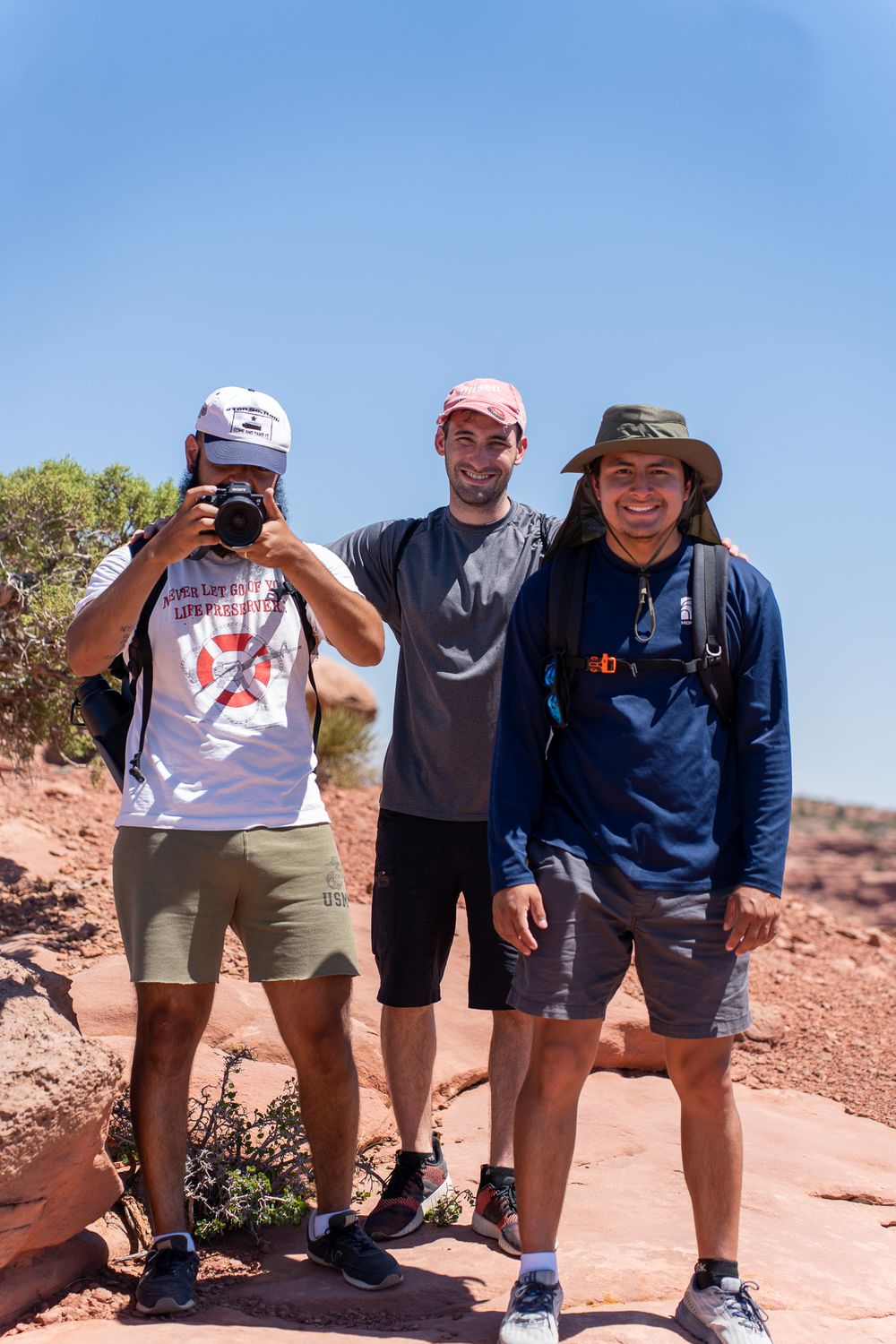 Three hikers pose on red rock terrain with camera gear and outdoor equipment against a bright blue sky.