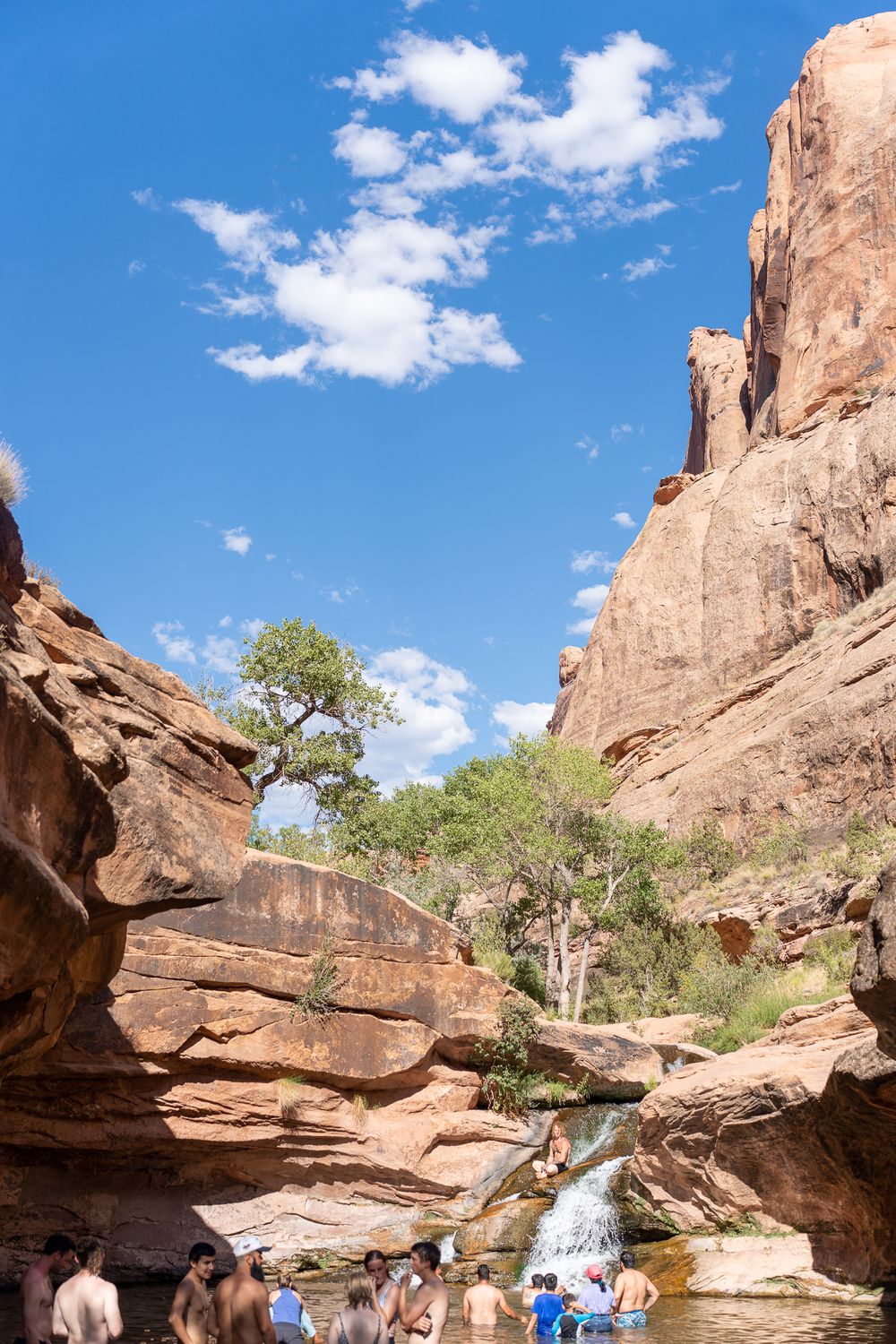 Visitors gather at a scenic waterfall oasis surrounded by towering red rock canyon walls.