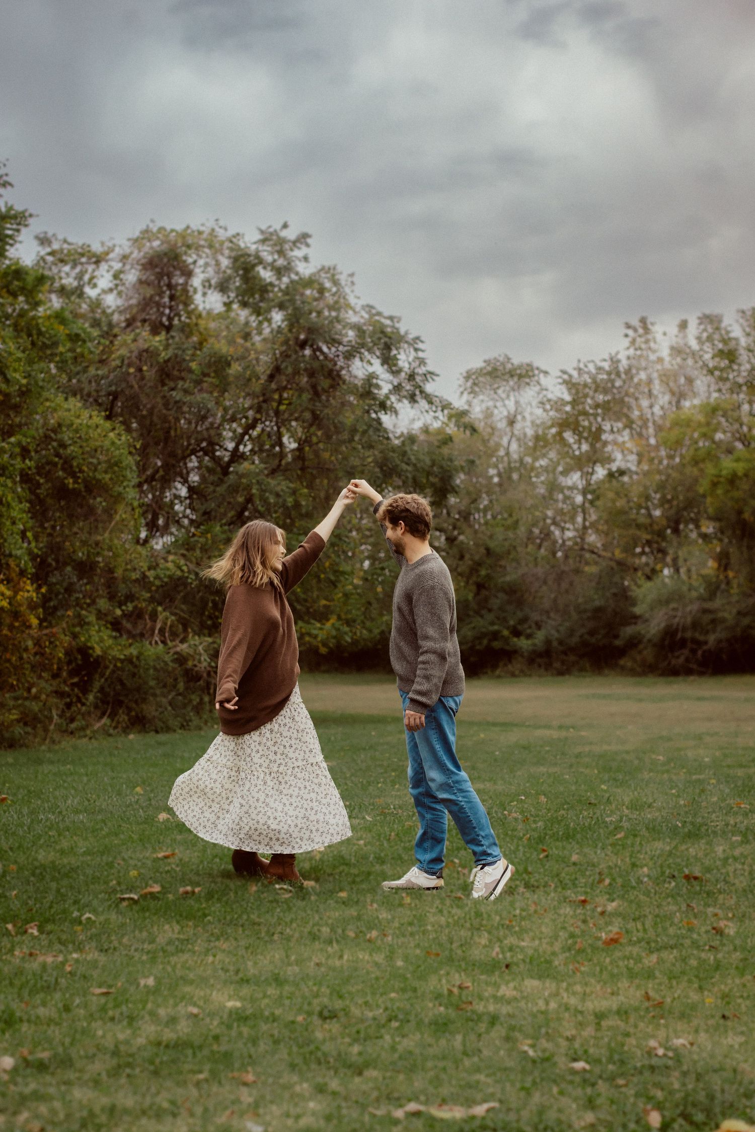 A man twirls a woman under a stormy sky during a dramatic, moody Kansas City engagement photo session.