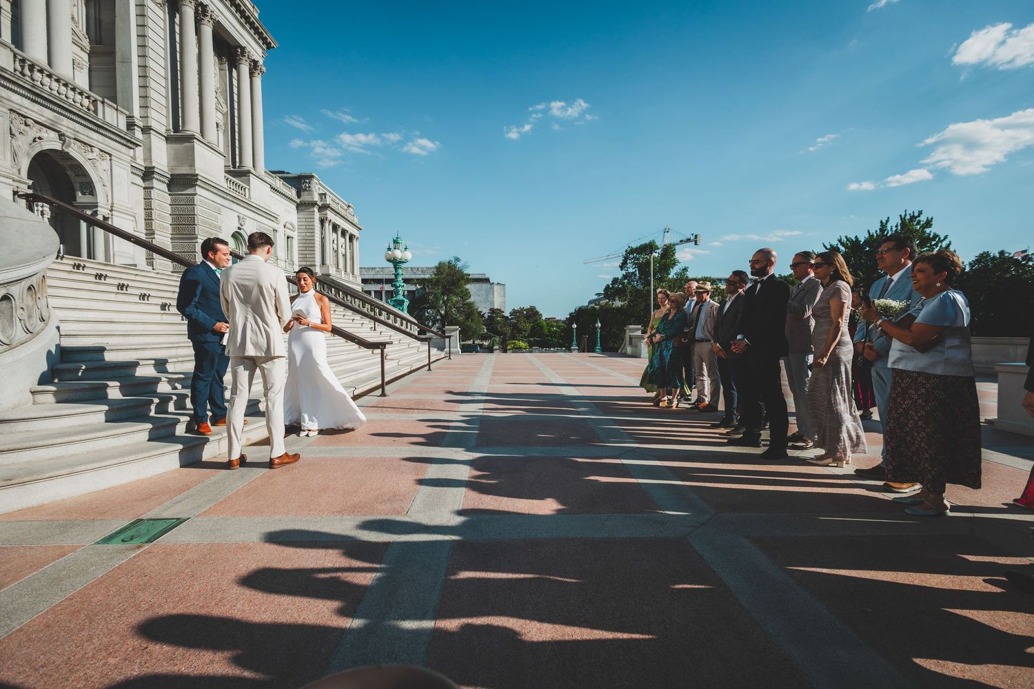 Wedding guests gather on steps of historic building as ceremony takes place under blue skies.