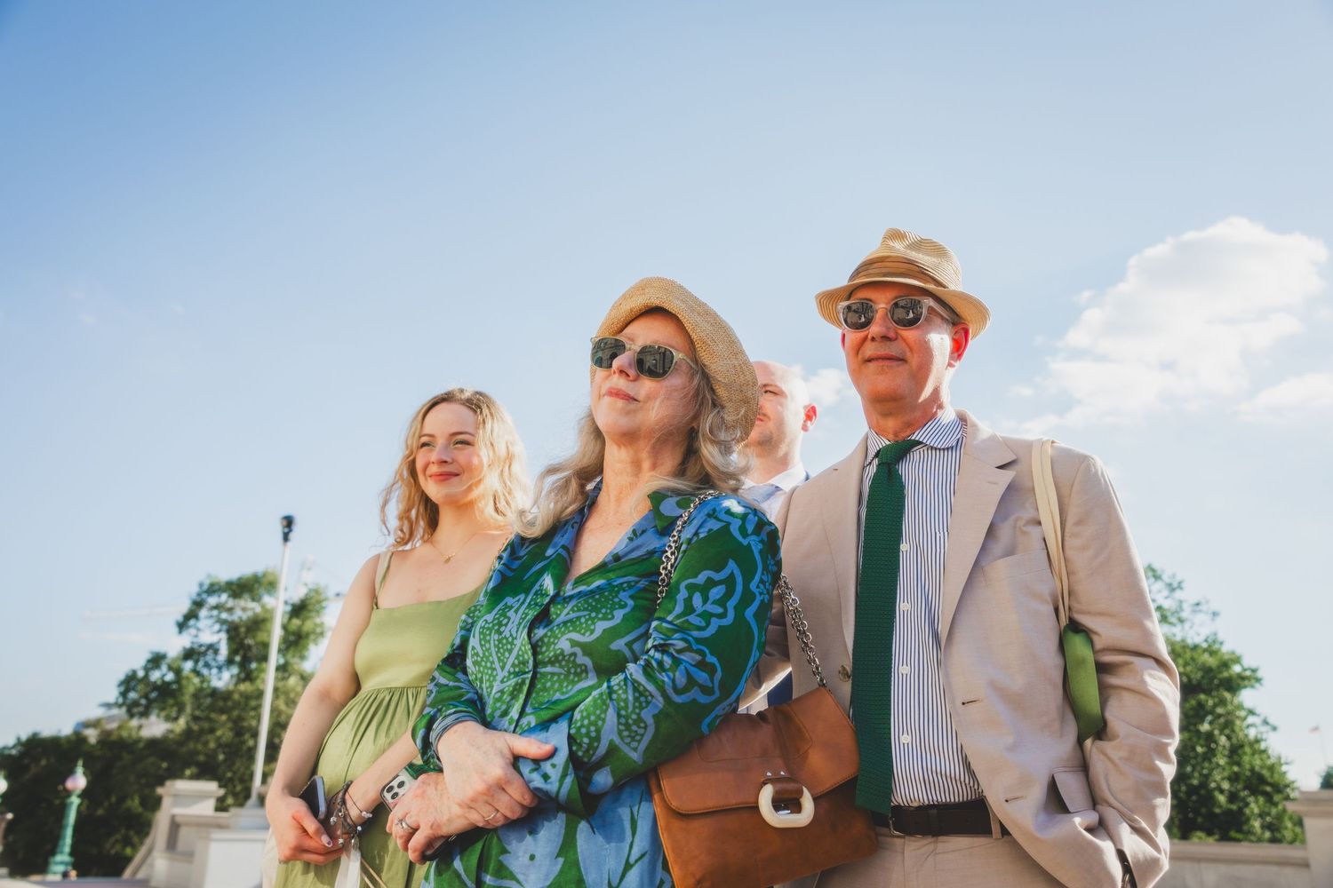 Wedding guests in summer attire pose on steps of historic building under bright blue sky.