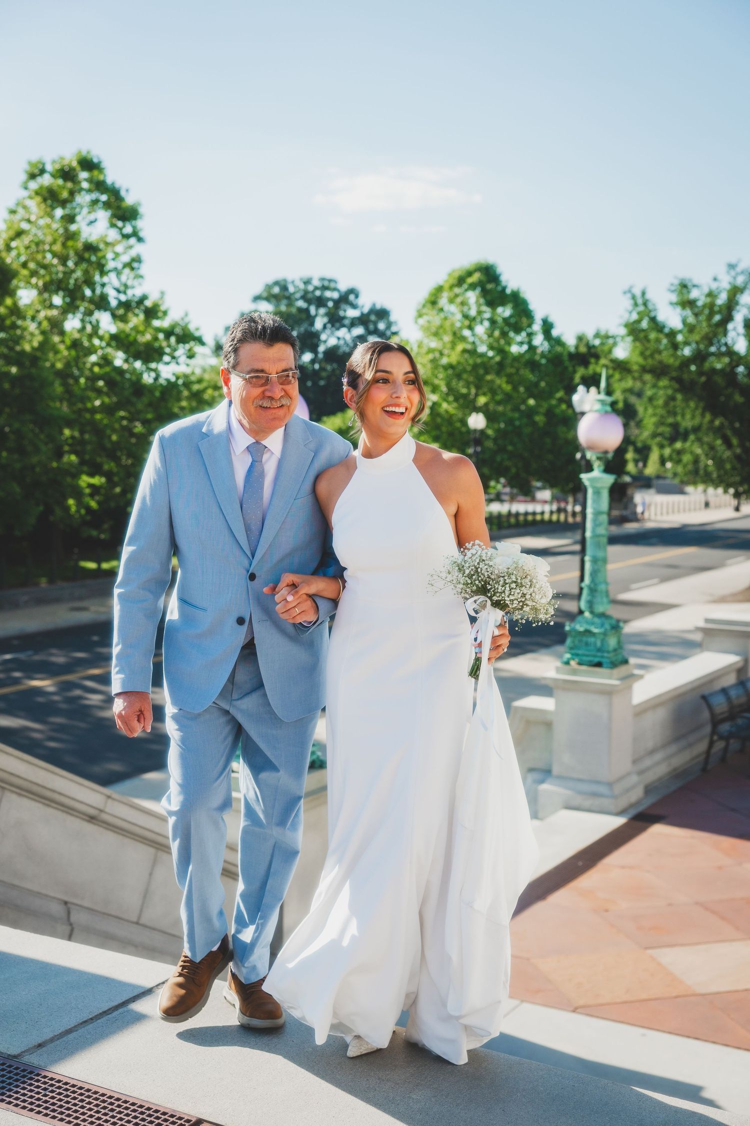 A couple poses elegantly on outdoor steps, with the person in a light blue suit escorting someone in a flowing white dress.