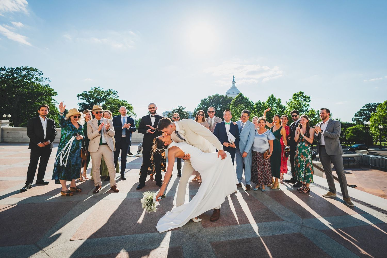 Wedding party celebrates with dramatic dip pose on sunny steps with Capitol dome in distance.