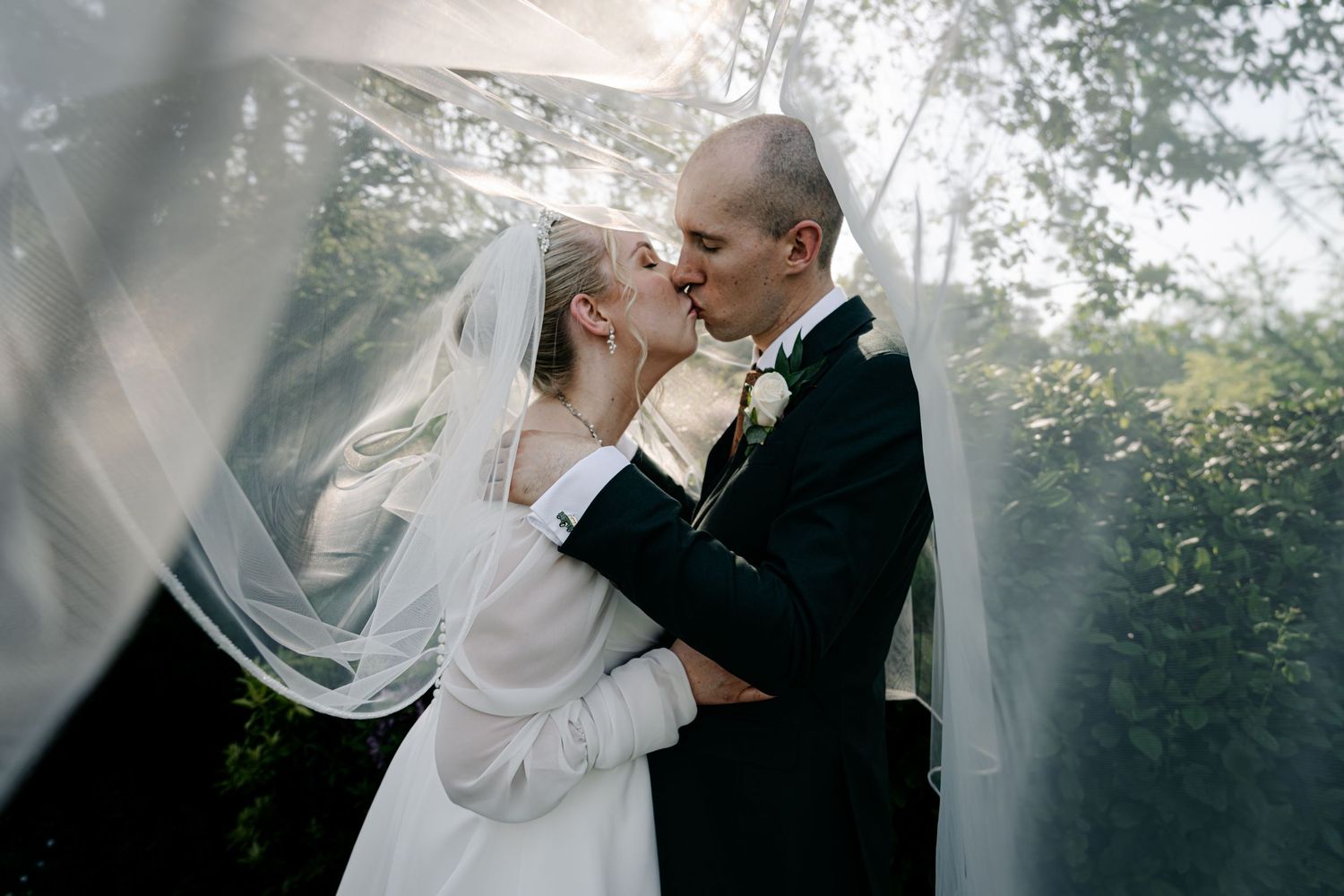 Two newlyweds share a romantic wedding kiss under a flowing white veil in soft lighting.