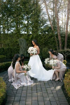 Bride in white gown holds bouquet as bridesmaids in taupe dresses arrange her train on garden stone path.
