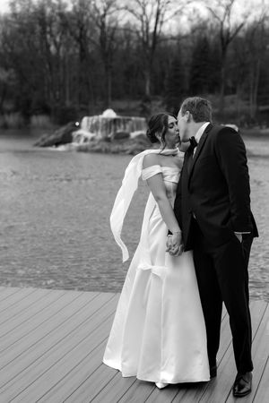 Black and white photo of bride and groom kissing on a dock near a lake with a fountain backdrop.