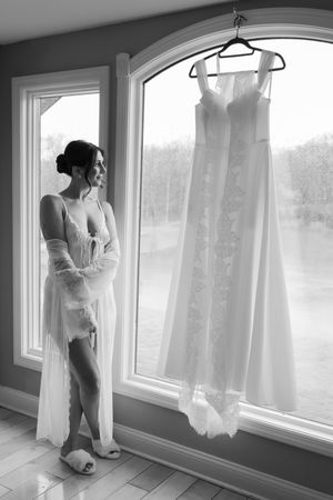 Bride in lingerie and sheer robe gazing at her wedding dress hanging by a large arched window.