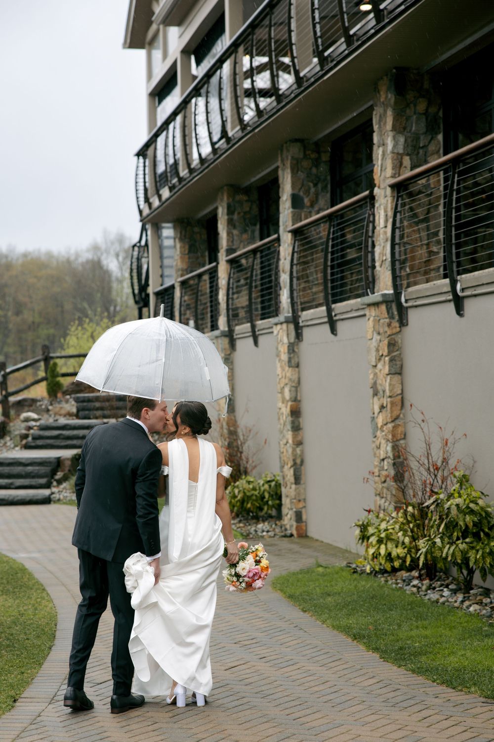 Bride and groom kissing under a white umbrella on a rainy wedding day outside a stone building.