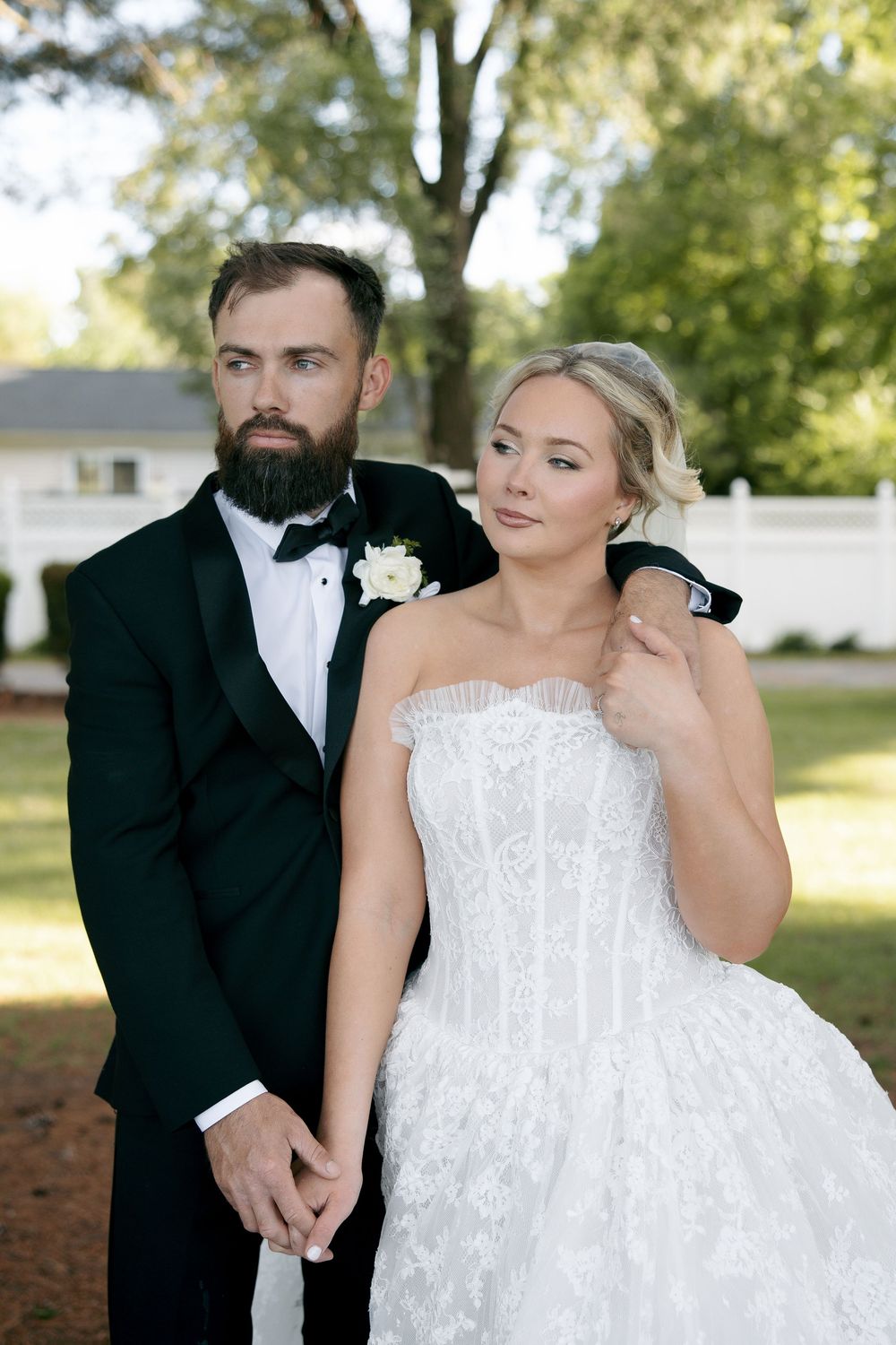Bride in strapless lace gown holds hands with bearded groom in black tuxedo outdoors at elegant wedding venue.