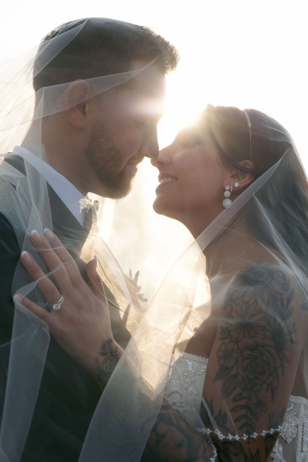 Bride and groom share romantic moment under veil with golden backlight, showcasing tattoos and wedding rings.